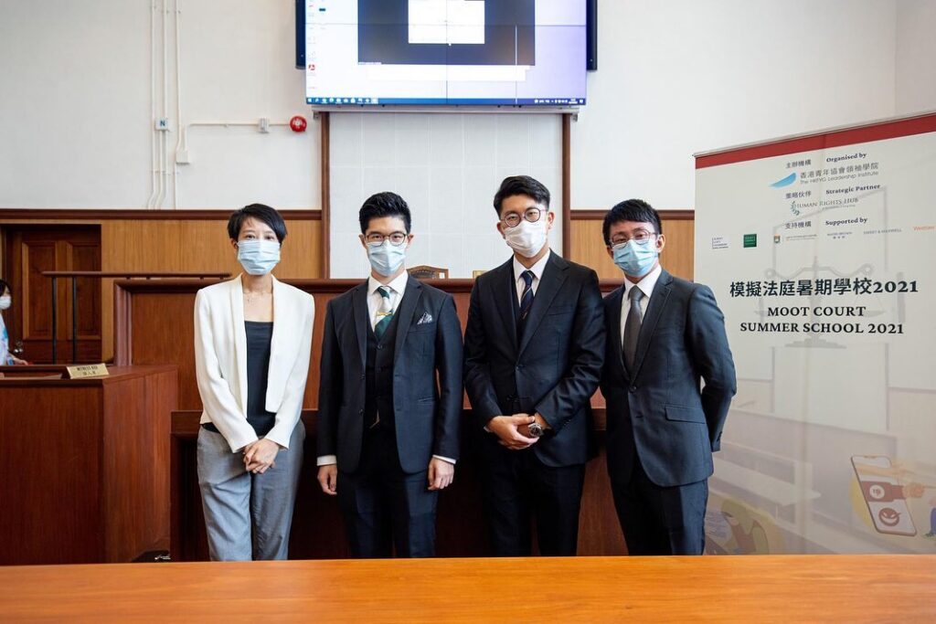 Four individuals posing in a courtroom, all wearing masks and formal attire, standing in front of a projector screen displaying legal text.