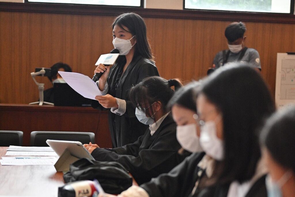 A young woman wearing a mask stands reading from a paper in a conference room filled with masked attendees during a community legal education session.