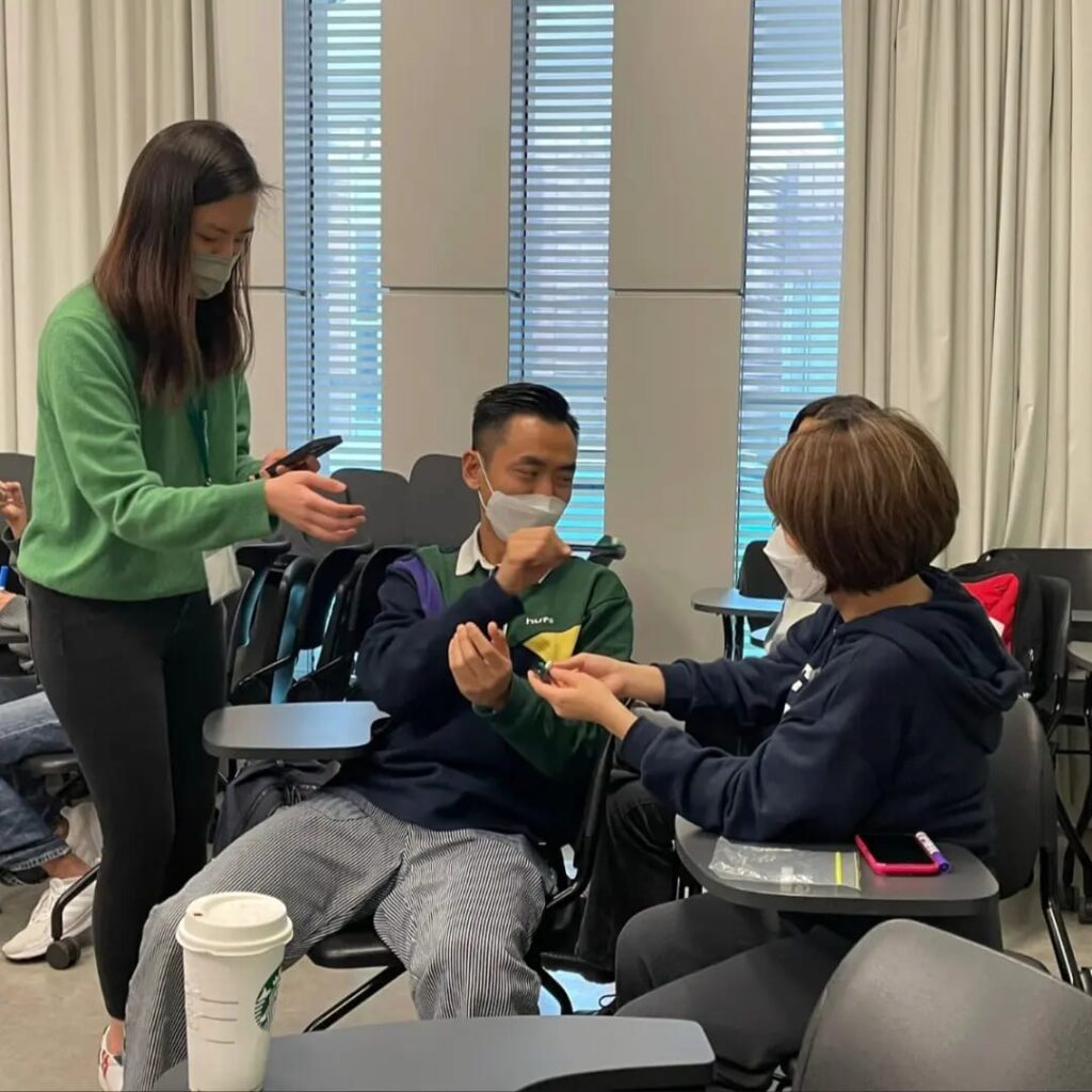 Three children in a classroom setting, exchanging cards while wearing masks. Two are seated, one stands, holding a smartphone.