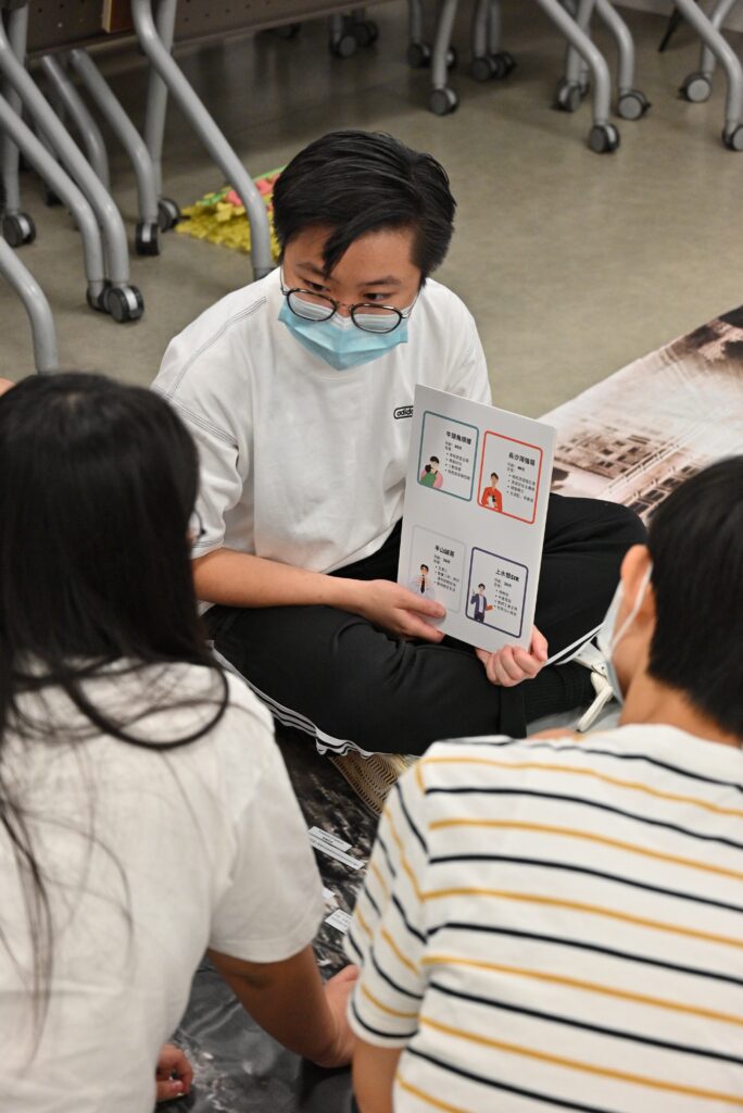 Person in mask holds open booklet showing legal diagrams, sitting on the floor while discussing with two others in a casual setting.