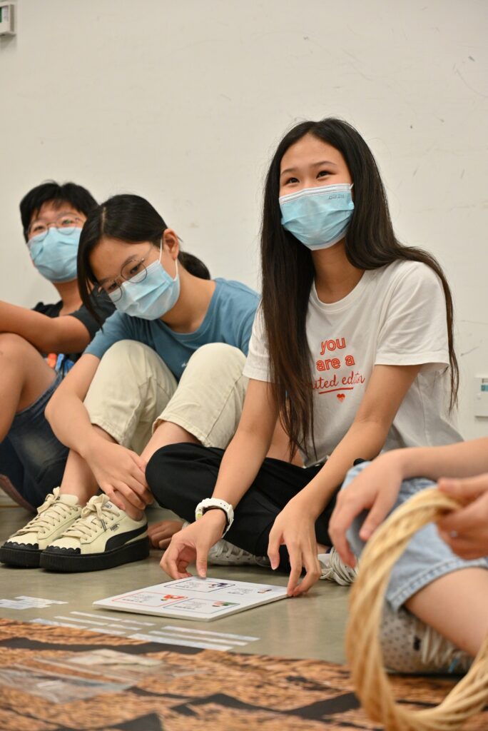 A group of young children wearing face masks sits on the floor, engaging in an educational activity with papers in front of them.