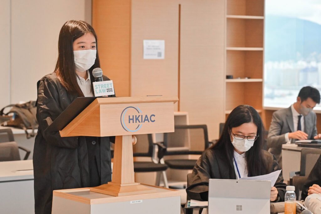 A woman in a mask speaks at a podium labeled "hkiac" during a law conference, while another attendee seated at a table looks at documents.