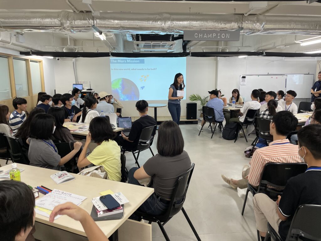 A woman presents to an audience in a workshop setting, with listeners seated at tables, focusing on a projector screen displaying "the mars mission," as part of a community legal education seminar on space law.