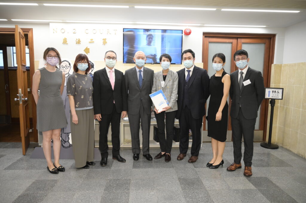 A group of eight individuals, wearing business attire and face masks, stands in a hallway outside a courtroom, holding law documents.