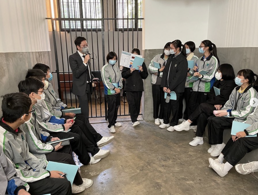 Man in suit speaking into microphone to group of children in masks sitting in semi-circle, in a room with barred windows, part of a community legal education program.