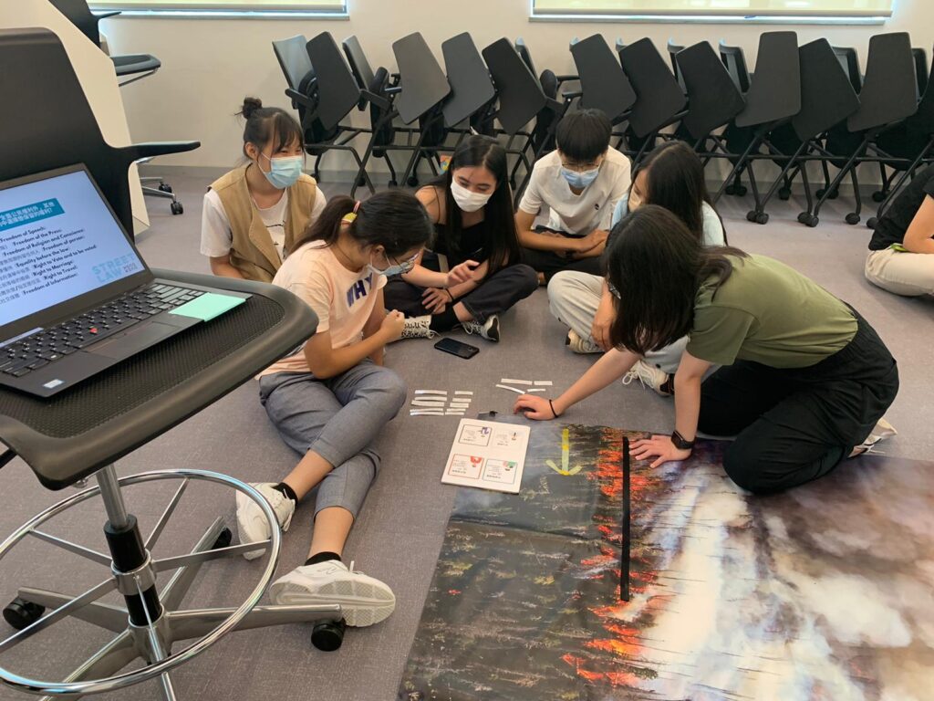 A group of young students, wearing face masks, sitting on the floor and engaging with educational materials in a community legal education classroom.